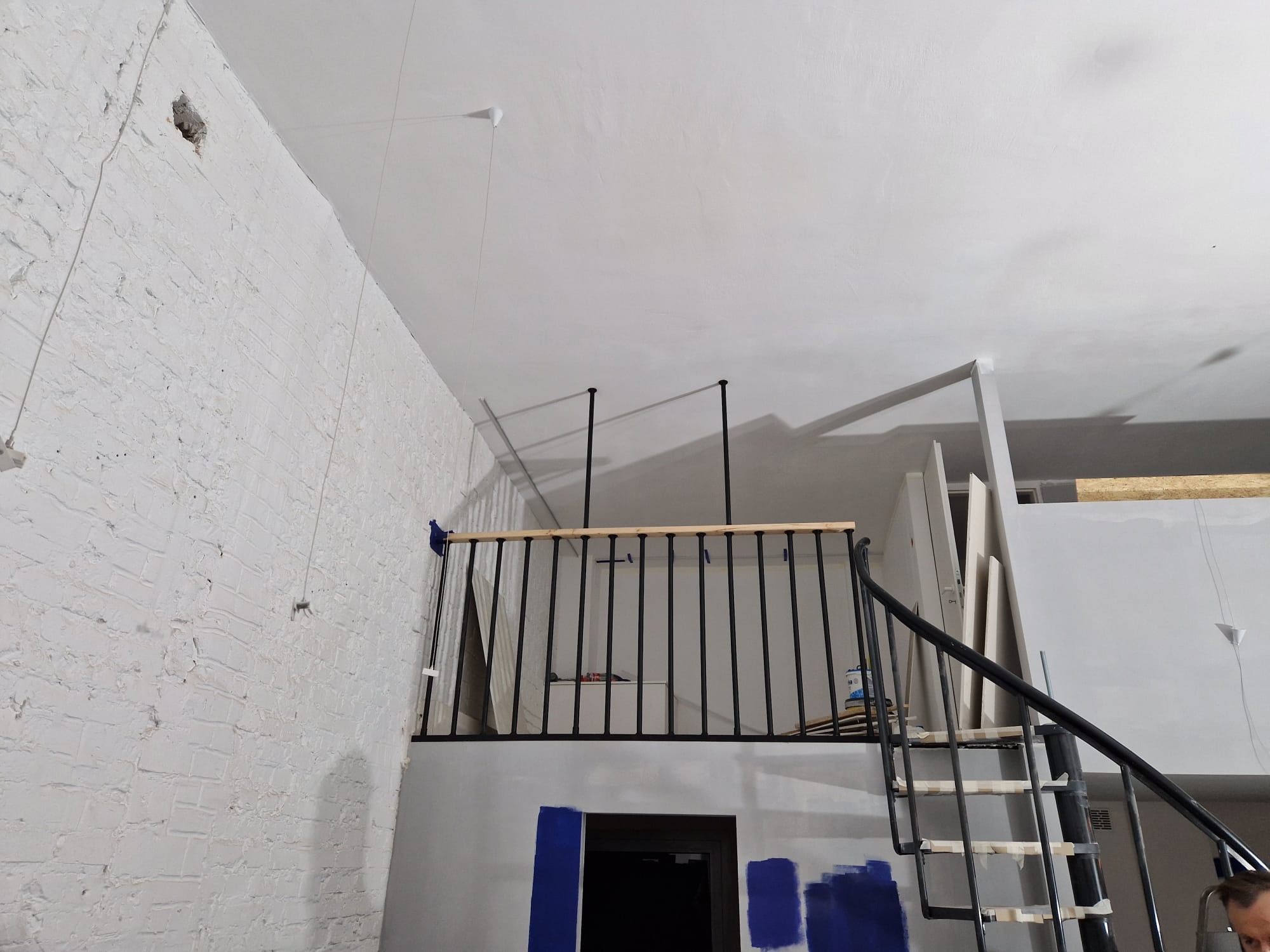 Wnętrze w trakcie remontu: biała ceglana ściana, czarna spiralna klatka schodowa, drewniana balustrada antresoli, ślady niebieskiej farby na ścianie, widoczne materiały budowlane.