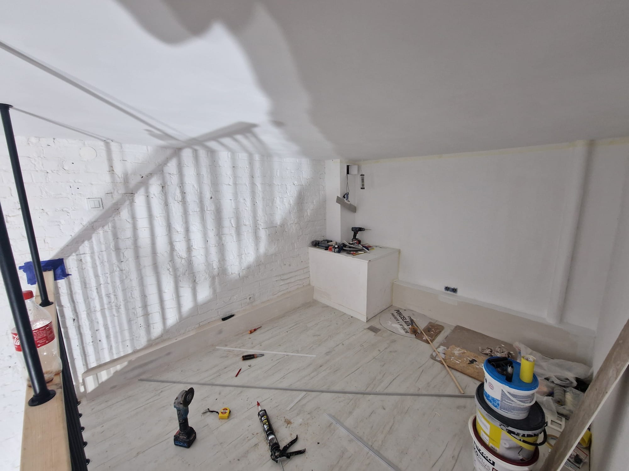 Wnętrze w trakcie remontu: biała ceglana ściana, świeżo pomalowana gładka ściana, narzędzia, puszki z farbą, listwy przypodłogowe i drewniane schody z czarną balustradą.