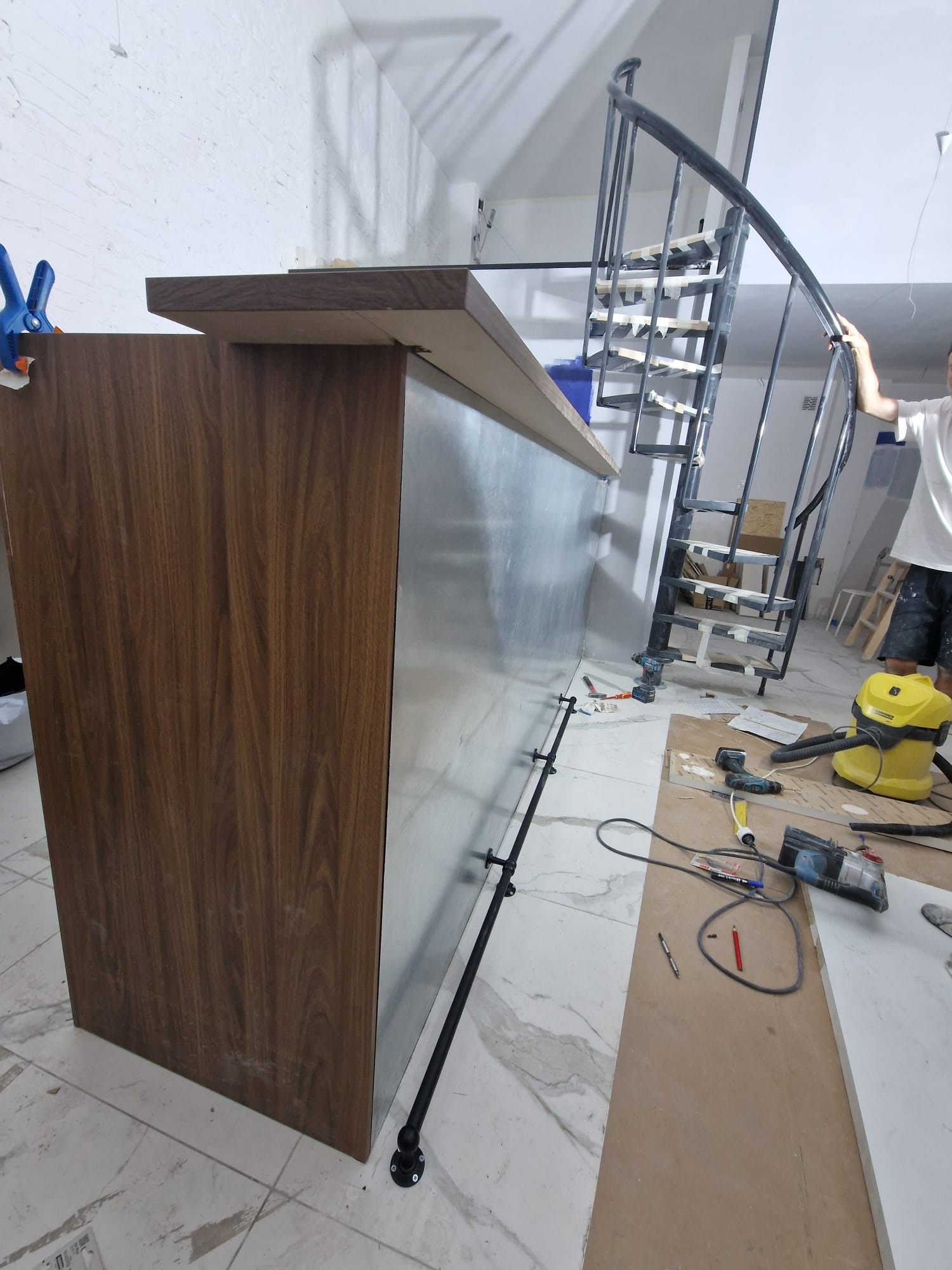 Montaż blatu barowego z drewnianym frontem i metalowym wykończeniem, widoczne narzędzia, odkurzacz i schody spiralne w tle.