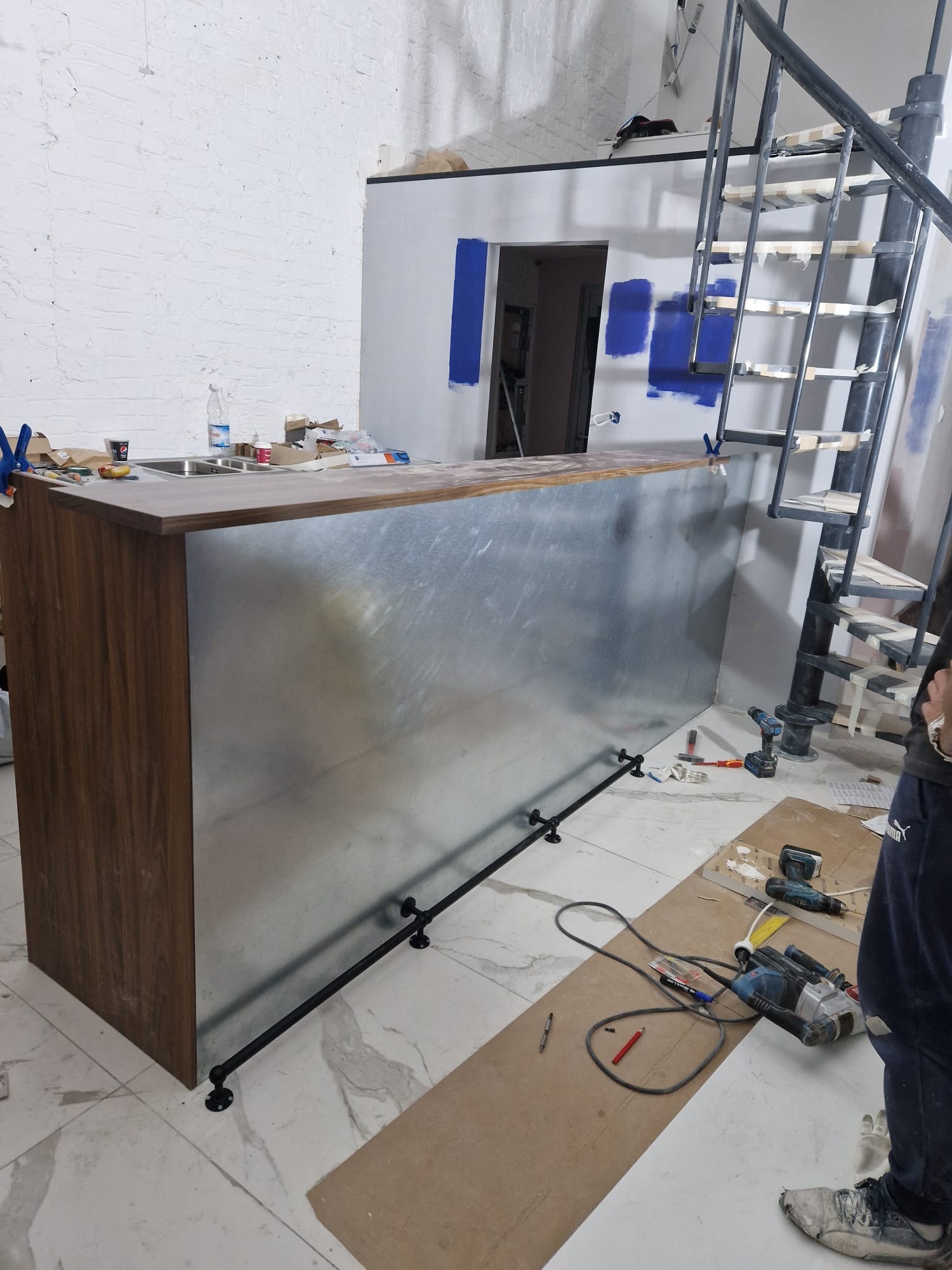 Wnętrze w trakcie remontu: metalowa lada barowa z drewnianym blatem, czarne rury dekoracyjne u dołu, narzędzia na podłodze, spiralne schody w tle, ślady malowania na ścianie.