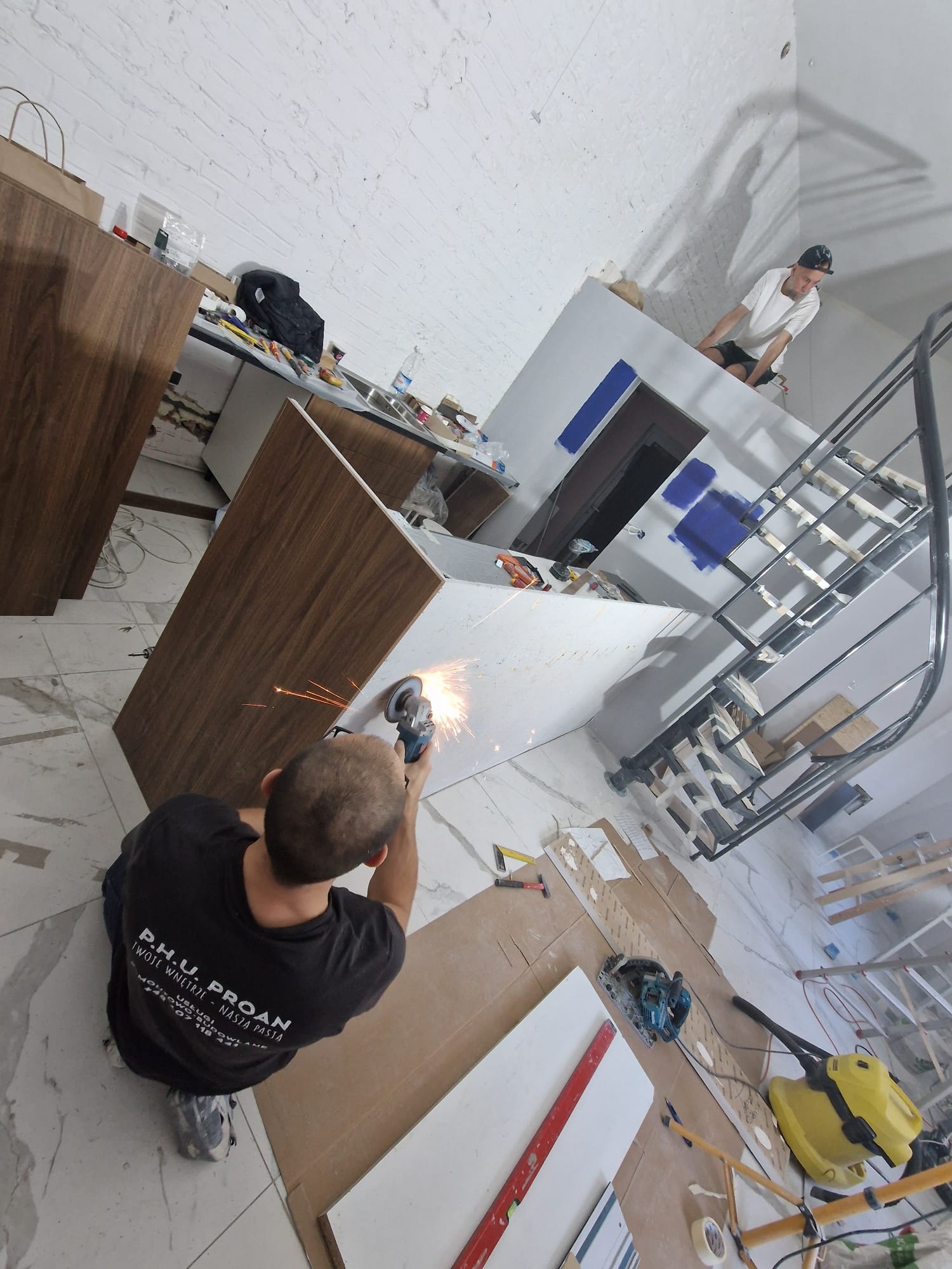 Montera szlifuje element meblowy szlifierką kątową, iskry lecą, w tle widoczne elementy zabudowy kuchennej i schody spiralne, na podłodze narzędzia i płyty