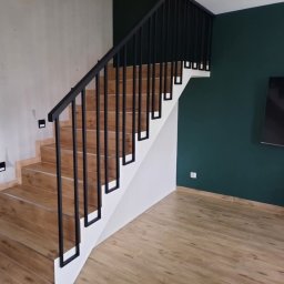 Steel Creative Mateusz Bartczak - Schody drewnopodobne z czarną, metalową balustradą o minimalistycznym designie, kontrastujące z ciemnozieloną ścianą w tle.