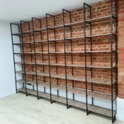 Duży, nowoczesny regał z czarną metalową ramą i drewnianymi półkami, ustawiony na tle ściany z czerwonej cegły, oświetlony z boku.