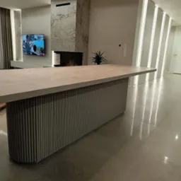 Duży, beżowy st&oacute;ł z blatem o nieregularnym kształcie i podstawą z pionowych, żebrowanych element&oacute;w, umieszczony w nowoczesnym salonie z marmurowym kominkiem i telewizorem.