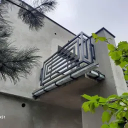 Nowoczesna balustrada balkonowa z czarnych profili o geometrycznym wzorze, zamontowana na betonowym balkonie, widok z dołu, z elementami zieleni w kadrze.