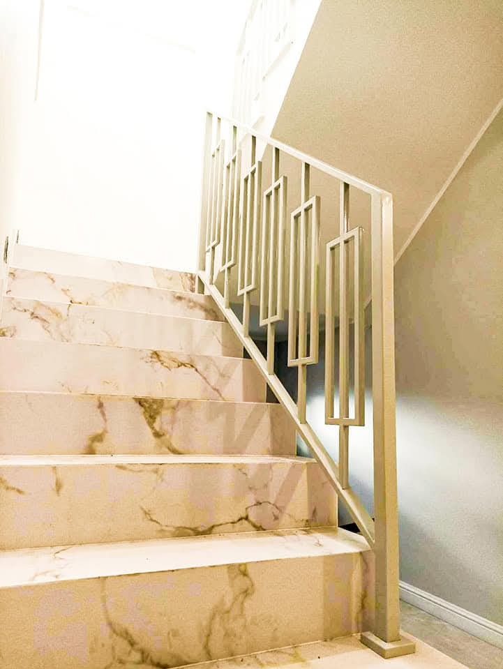 Schody wyłożone marmurem z nowoczesną, metalową balustradą w Ostrowie Wielkopolskim. Balustrada o geometrycznym wzorze, malowana na jasny kolor, kontrastuje z ciepłym odcieniem marmuru.