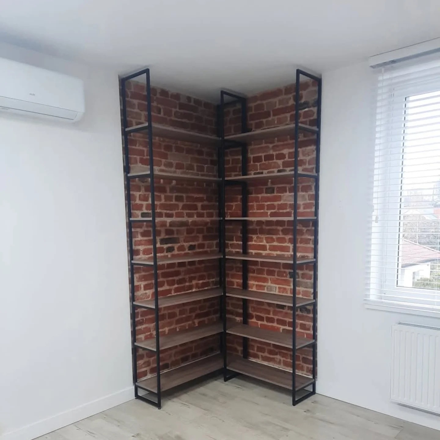 Kątowy regał z metalową ramą i półkami z płyty wiórowej, umieszczony na tle ściany z czerwonej cegły, z widocznym klimatyzatorem i oknem z żaluzjami.