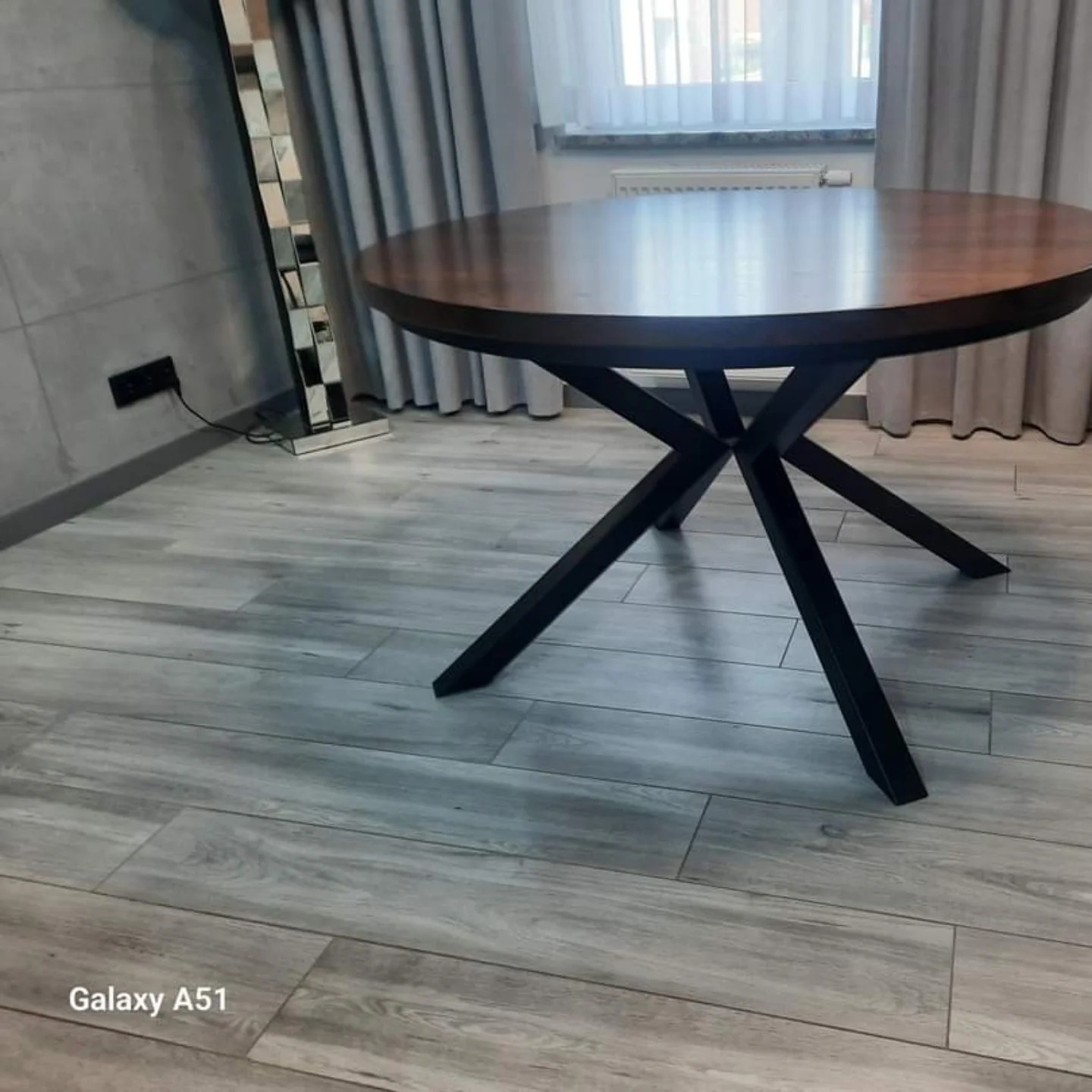 Okrągły stół z drewnianym blatem i metalową, krzyżową podstawą w nowoczesnym wnętrzu z szarymi panelami ściennymi i zasłonami.