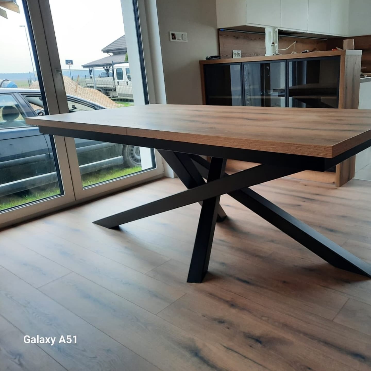 Stół z drewnianym blatem i czarnymi, metalowymi nogami w kształcie litery X, widoczny w jasnym wnętrzu z dużym oknem i drewnianą podłogą.