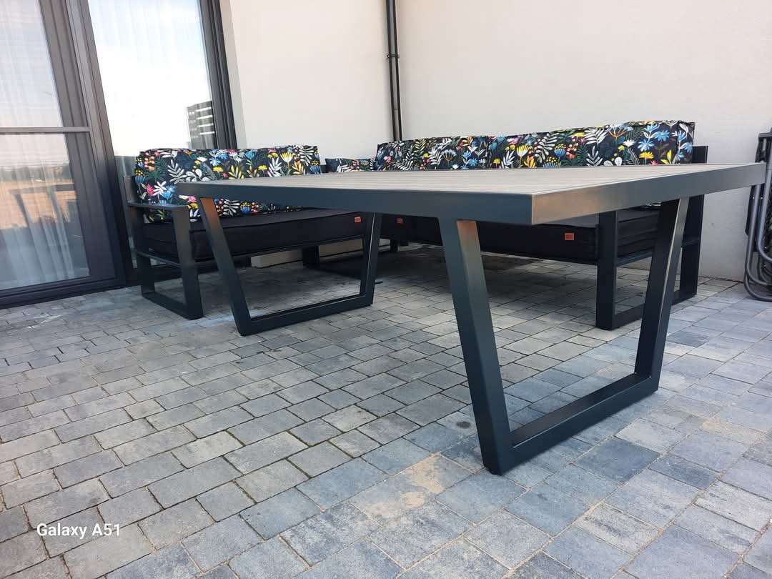 Zestaw mebli ogrodowych z metalową ramą i poduszkami w kwiatowe wzory, ustawiony na szarym brukowanym tarasie przylegającym do domu z dużymi przesuwnymi drzwiami.