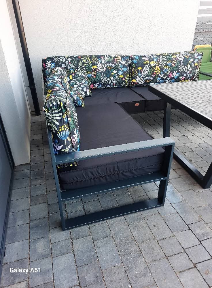 Metalowa konstrukcja sofy ogrodowej z czarnymi poduszkami i wzorzystymi poduchami w kwiaty i liście, stojąca na szarym bruku.