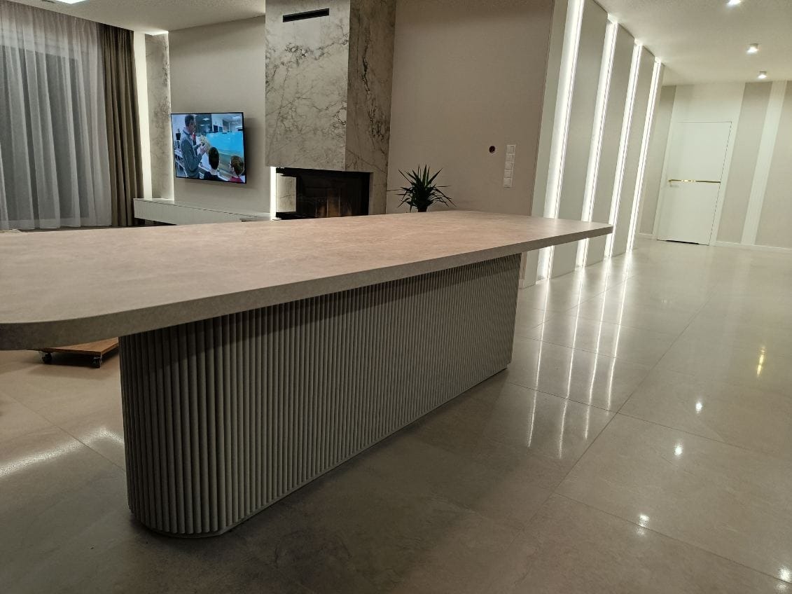 Duży, beżowy stół z blatem o nieregularnym kształcie i podstawą z pionowych, żebrowanych elementów, umieszczony w nowoczesnym salonie z marmurowym kominkiem i telewizorem.