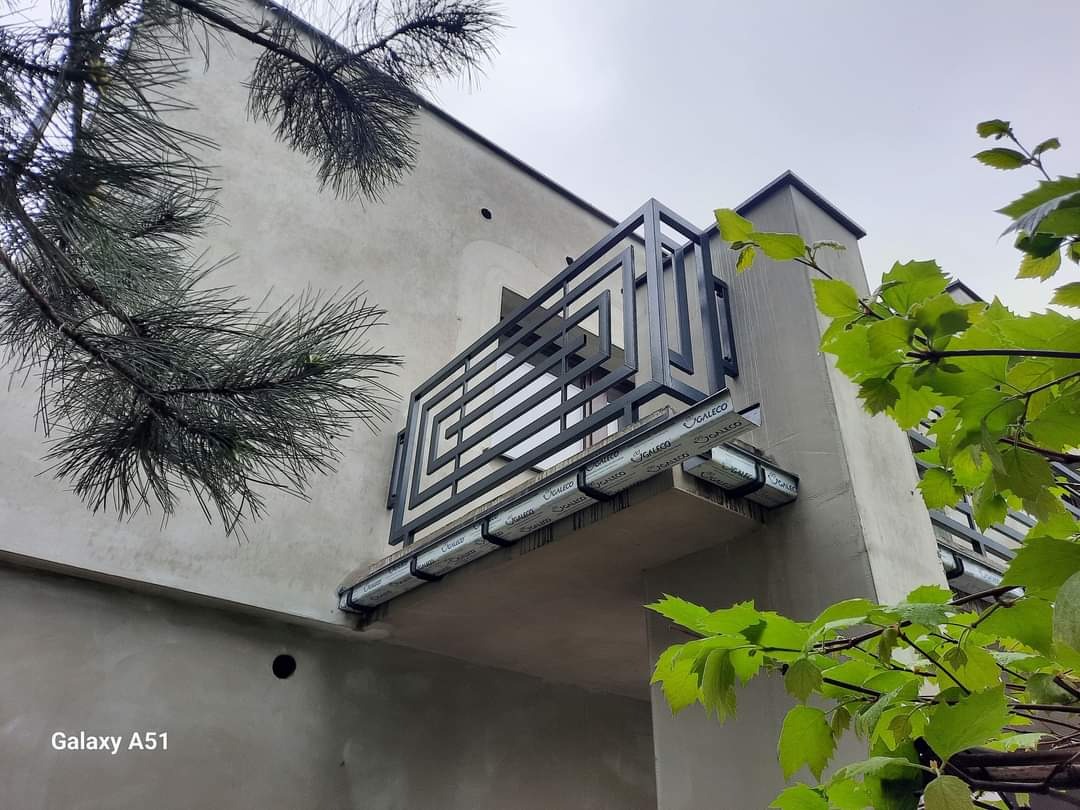 Nowoczesna balustrada balkonowa z czarnych profili o geometrycznym wzorze, zamontowana na betonowym balkonie, widok z dołu, z elementami zieleni w kadrze.