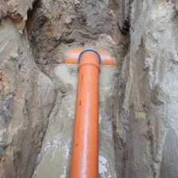 Pomarańczowa rura kanalizacyjna w wykopie wypełnionym wodą, widok z góry na połączenie trójnikowe.