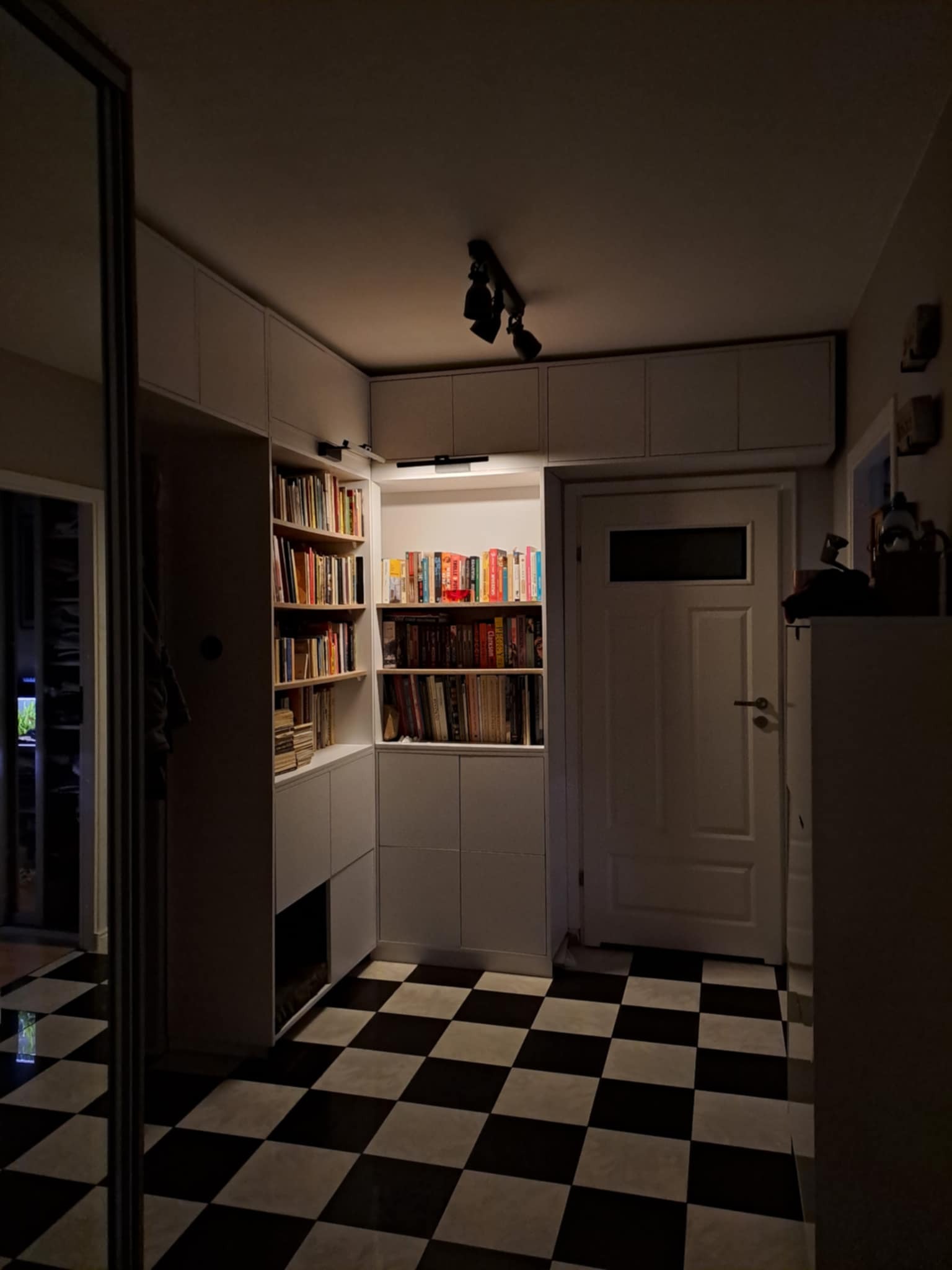 Biała szafa wnękowa z półkami na książki w przedpokoju z czarno-białą szachownicą na podłodze i oświetleniem punktowym na suficie.