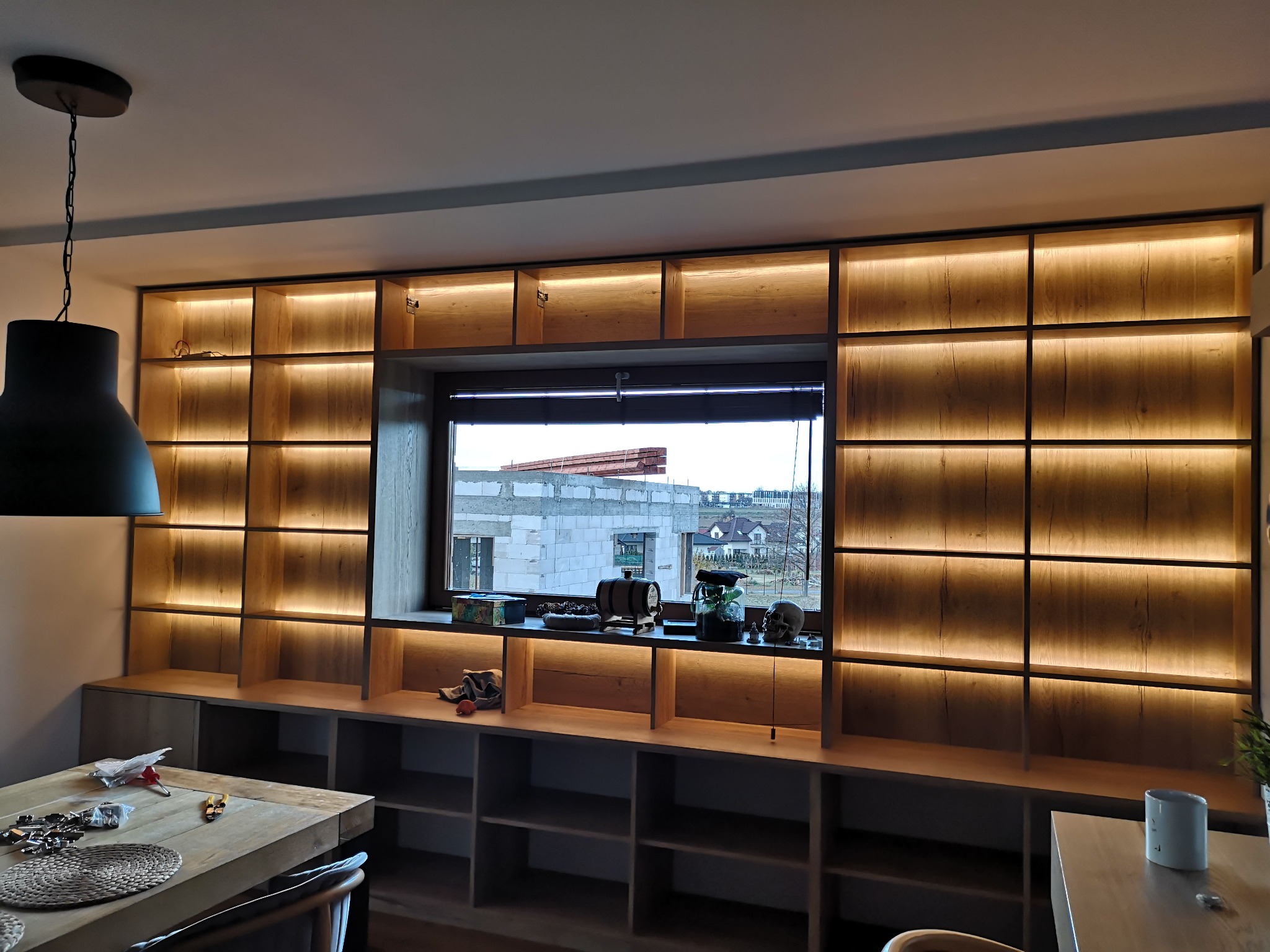 Drewniana zabudowa ściany z półkami i podświetleniem LED, widok na okno z elementami budowy na zewnątrz, stół z narzędziami na pierwszym planie.