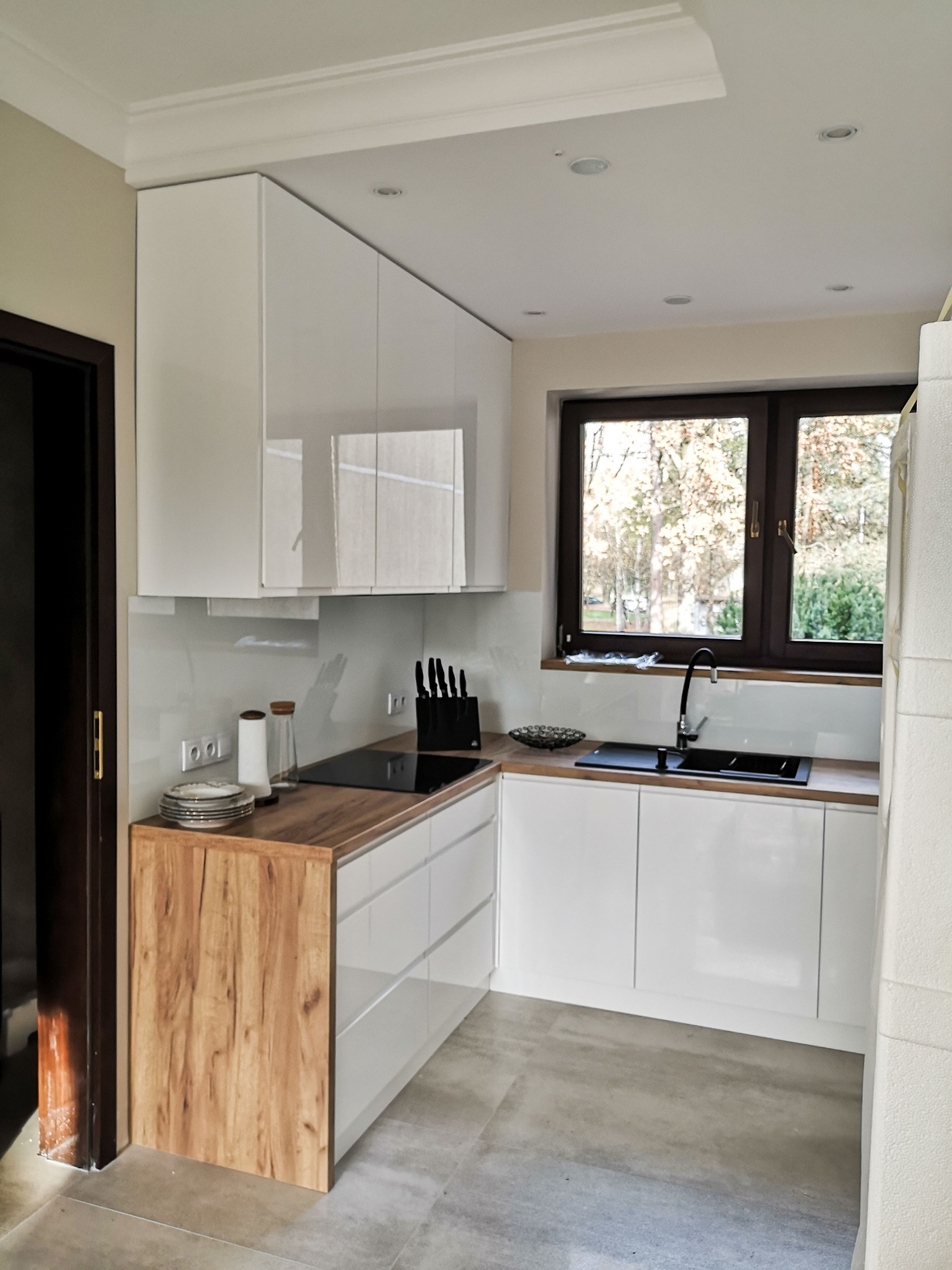 Nowoczesna kuchnia z białymi, lakierowanymi szafkami wiszącymi i dolnymi, drewnianym blatem, czarnym zlewem i płytą indukcyjną, widok na jesienny ogród przez okno.