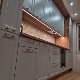 Meblopolis - Nowoczesna kuchnia z szarymi szafkami, drewnianymi akcentami i oświetleniem LED pod szafkami wiszącymi, na tle ściany z mozaiką.