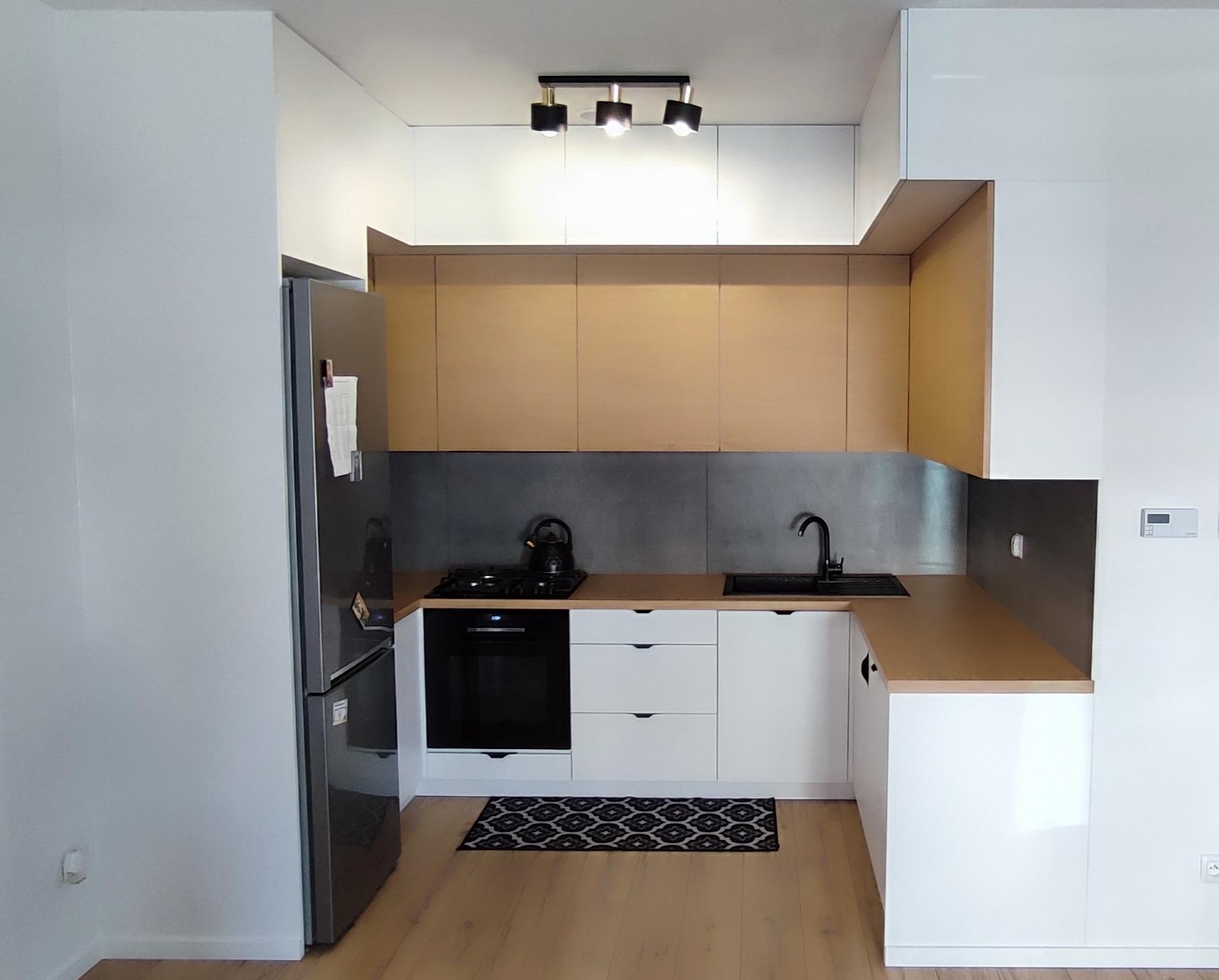 Nowoczesna kuchnia z białymi i drewnianymi szafkami, czarnym zlewozmywakiem i płytą grzewczą, oraz szarym panelem ściennym. Mała czarno-biała wycieraczka na podłodze.