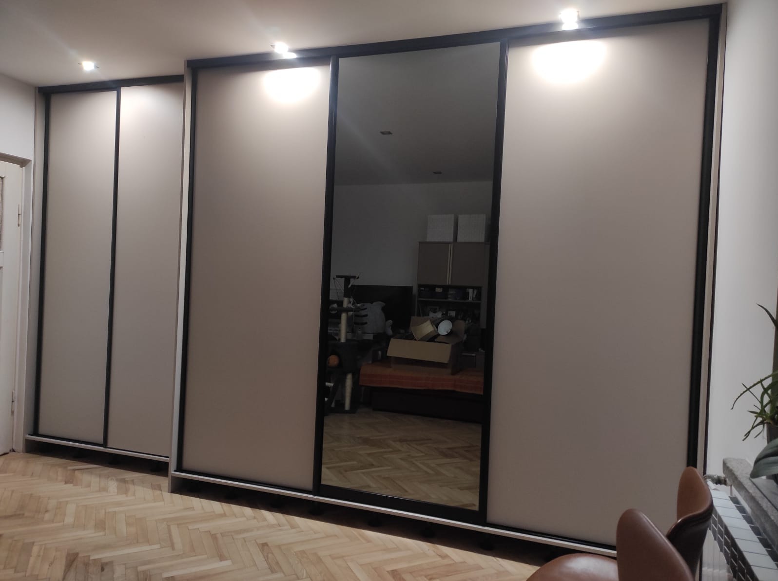 Szafa przesuwna z lustrem w czarnej ramie, odbijająca wnętrze pokoju z parkietem w jodełkę.