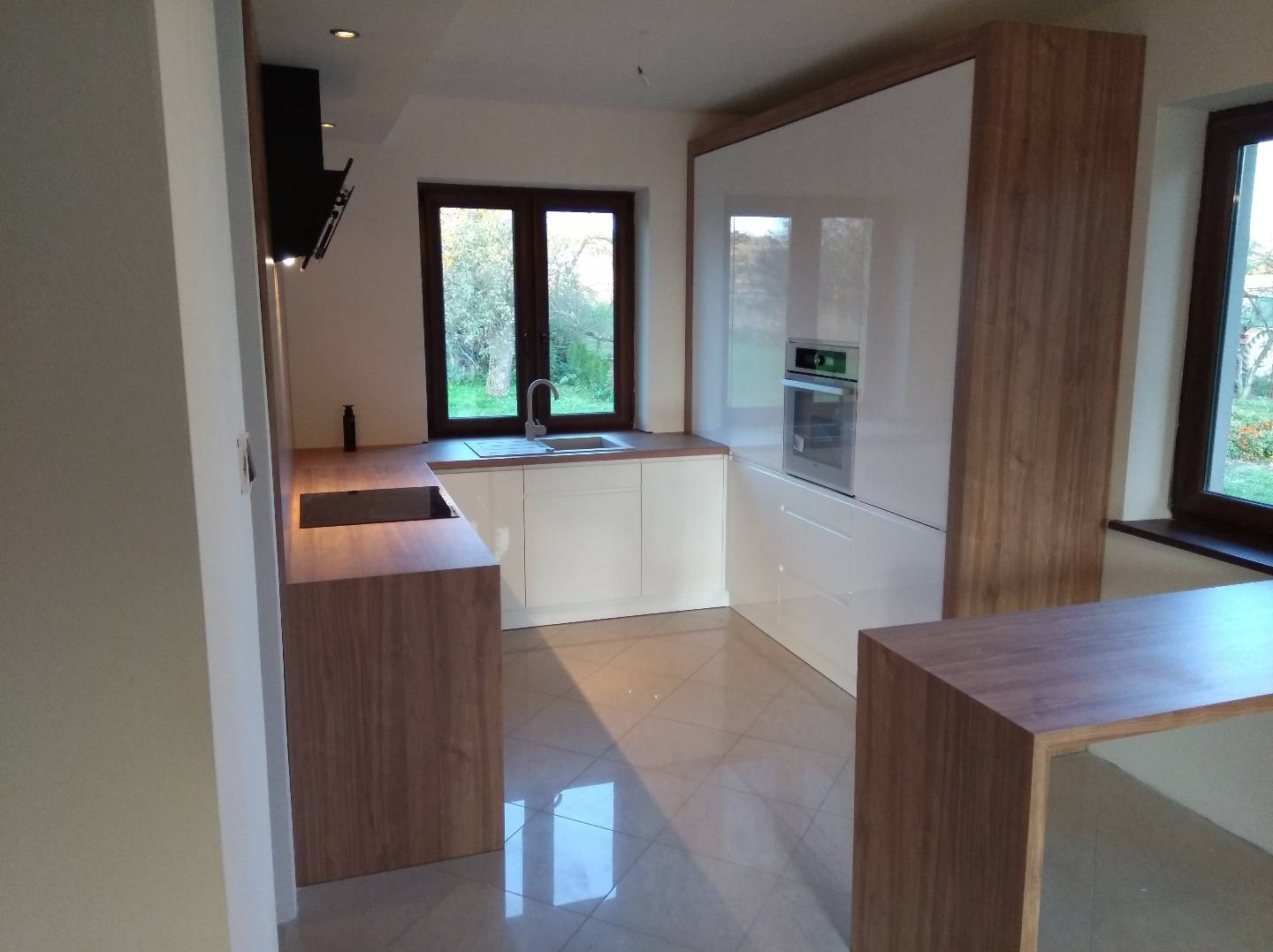 Nowoczesna kuchnia z białymi frontami i drewnianymi akcentami, widok na okno z ogrodem, płytki podłogowe z połyskiem.