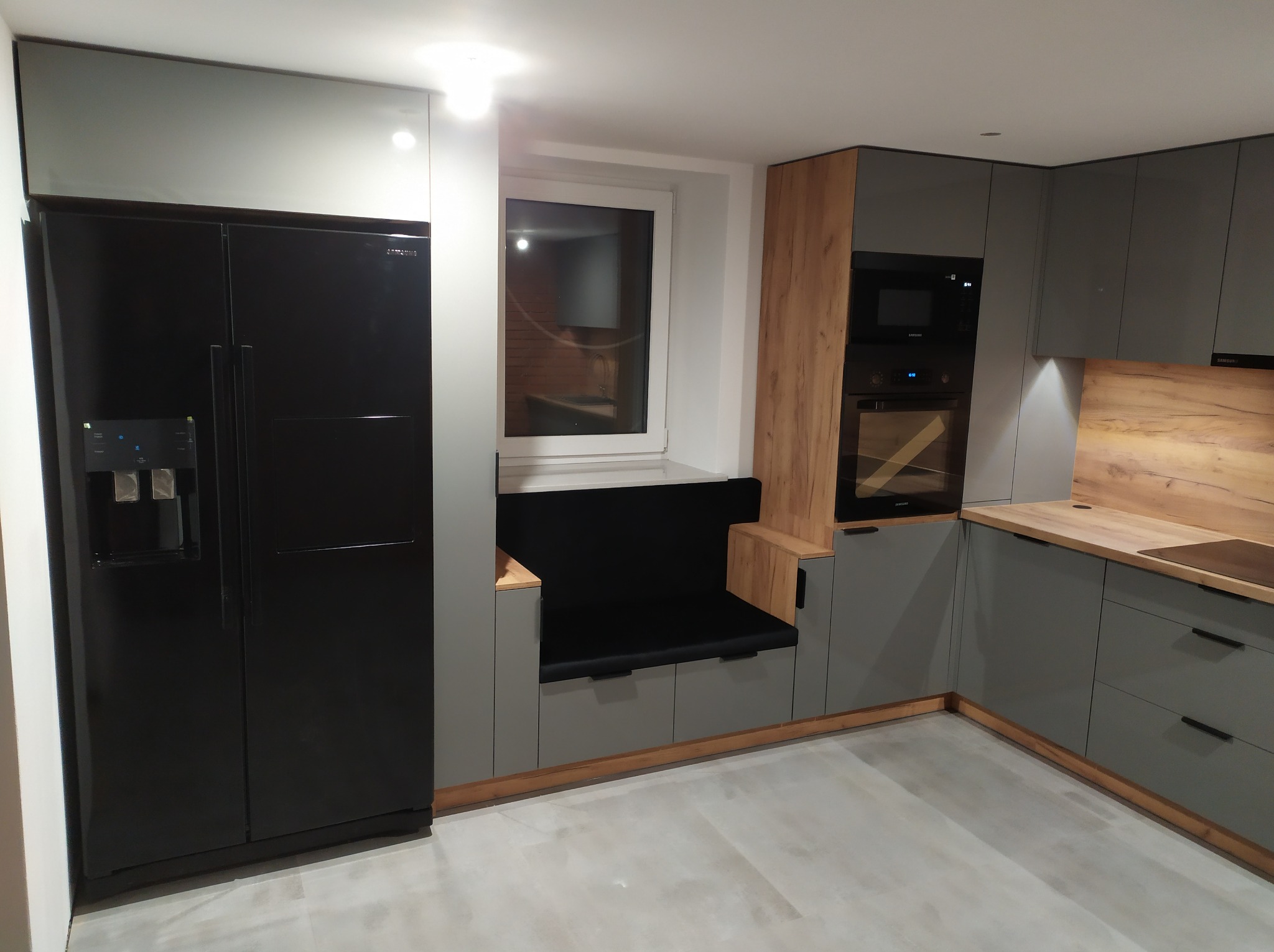 Nowoczesna kuchnia na wymiar z szarymi szafkami, drewnianymi akcentami i czarną lodówką Samsung z kostkarką, z wkomponowanym siedziskiem pod oknem.