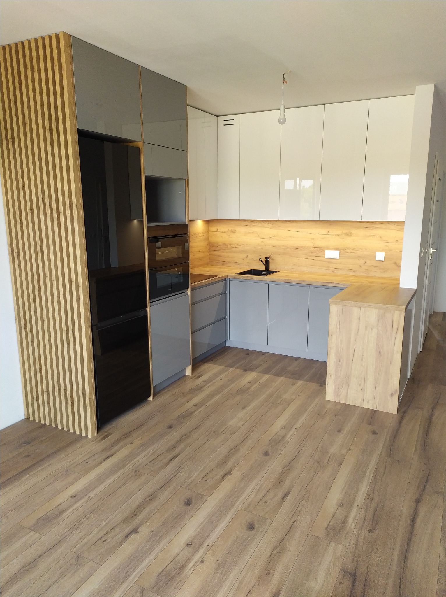 Nowoczesna kuchnia z szarymi i białymi szafkami, drewnianym blatem i pionowymi drewnianymi panelami dekoracyjnymi, czarny zlewozmywak, oświetlenie podszafkowe, podłoga z paneli drewnopodobnych.