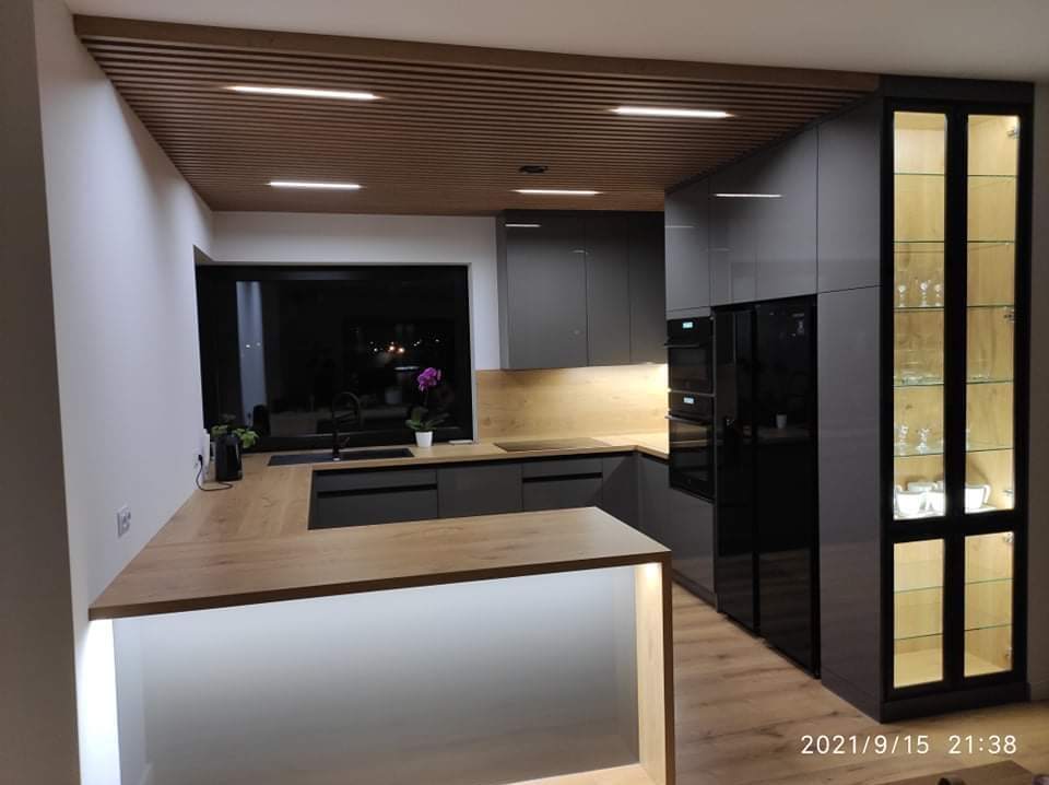 Nowoczesna kuchnia w odcieniach szarości z drewnianym blatem i podświetlanym barkiem, drewniany sufit lamelowy z oświetleniem punktowym, przeszklona witryna z oświetleniem wewnętrznym.