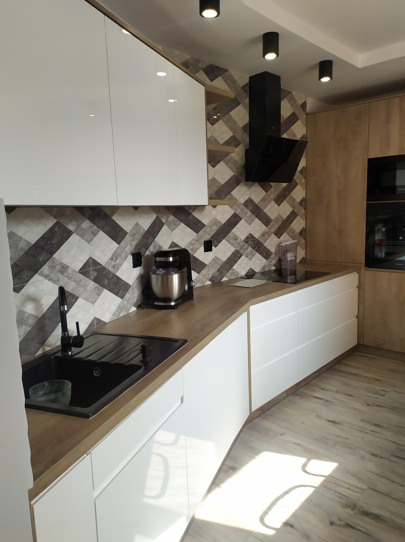 Nowoczesna kuchnia z białymi frontami i drewnianym blatem, czarny zlew i bateria, geometryczna mozaika na ścianie, czarny okap i sprzęt AGD.