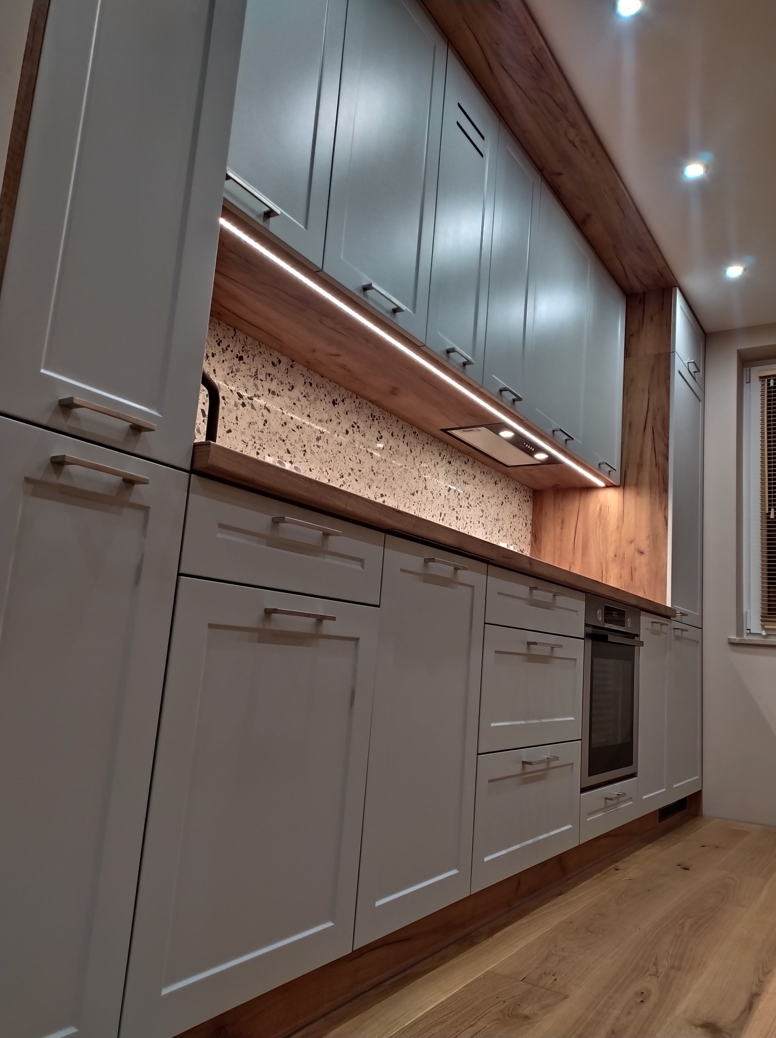 Nowoczesna kuchnia z szarymi szafkami, drewnianymi akcentami i oświetleniem LED pod szafkami wiszącymi, na tle ściany z mozaiką.