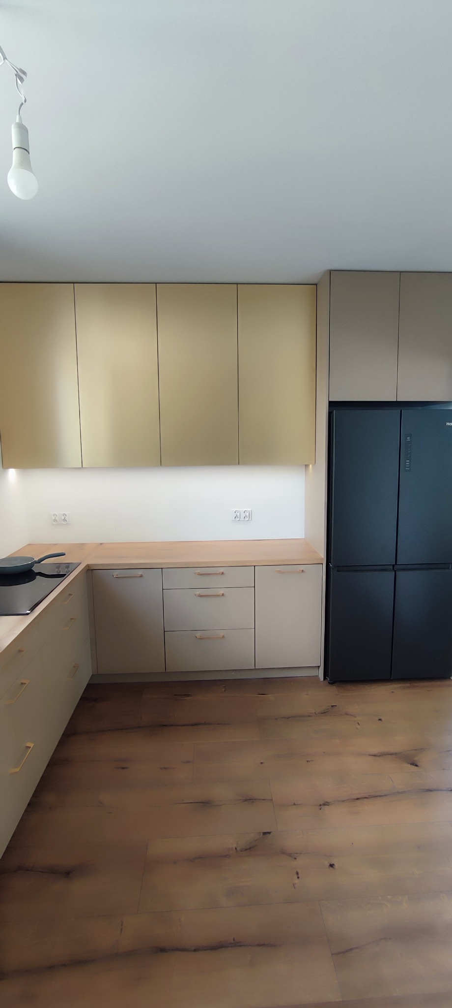 Nowoczesna kuchnia z górnymi szafkami w kolorze złota i dolnymi w odcieniach beżu, drewniany blat, czarna lodówka side-by-side, parkiet na podłodze.