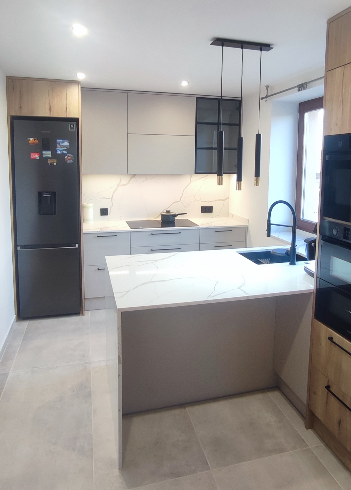 Nowoczesna kuchnia z wyspą, szare szafki z czarnymi uchwytami, biały blat z marmurowym wzorem, czarny zlew i bateria, czarna lodówka side-by-side, wiszące lampy cylindryczne z elementami złotymi.