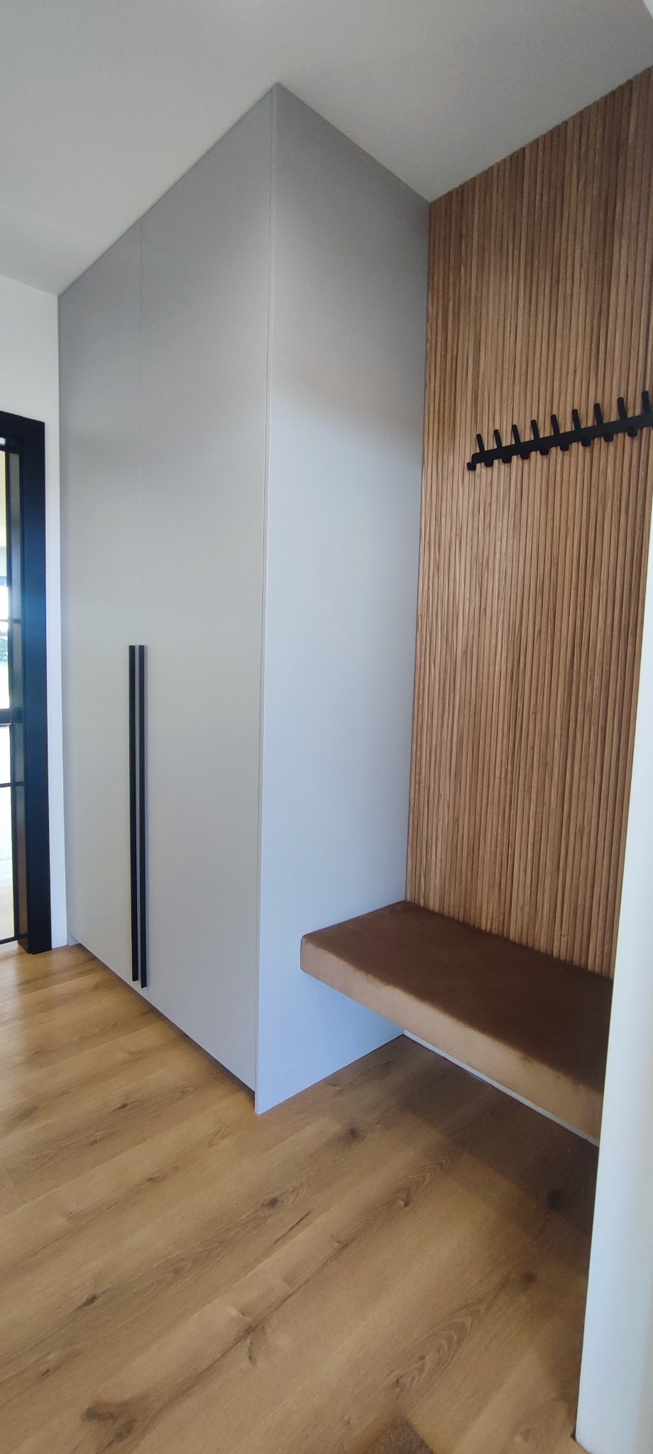 Szara szafa w przedpokoju z czarnymi uchwytami, drewniany panel ścienny z wieszakami i siedziskiem w kolorze brązowym, na podłodze panele imitujące drewno.