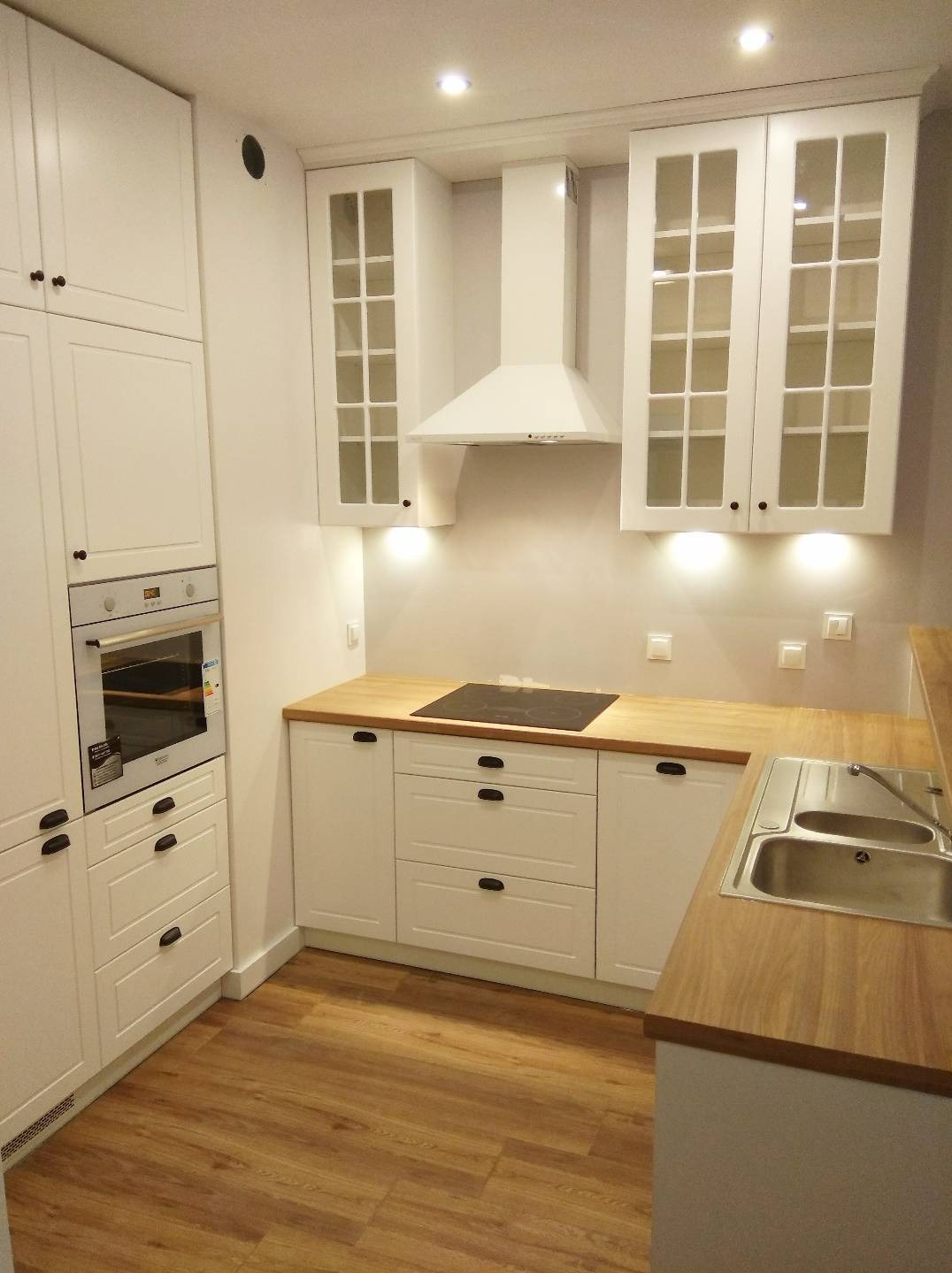 Jasna, nowoczesna zabudowa kuchenna z drewnianym blatem w Zamościu. Widoczne szklane witryny szafek wiszących, piekarnik w zabudowie i płyta indukcyjna.