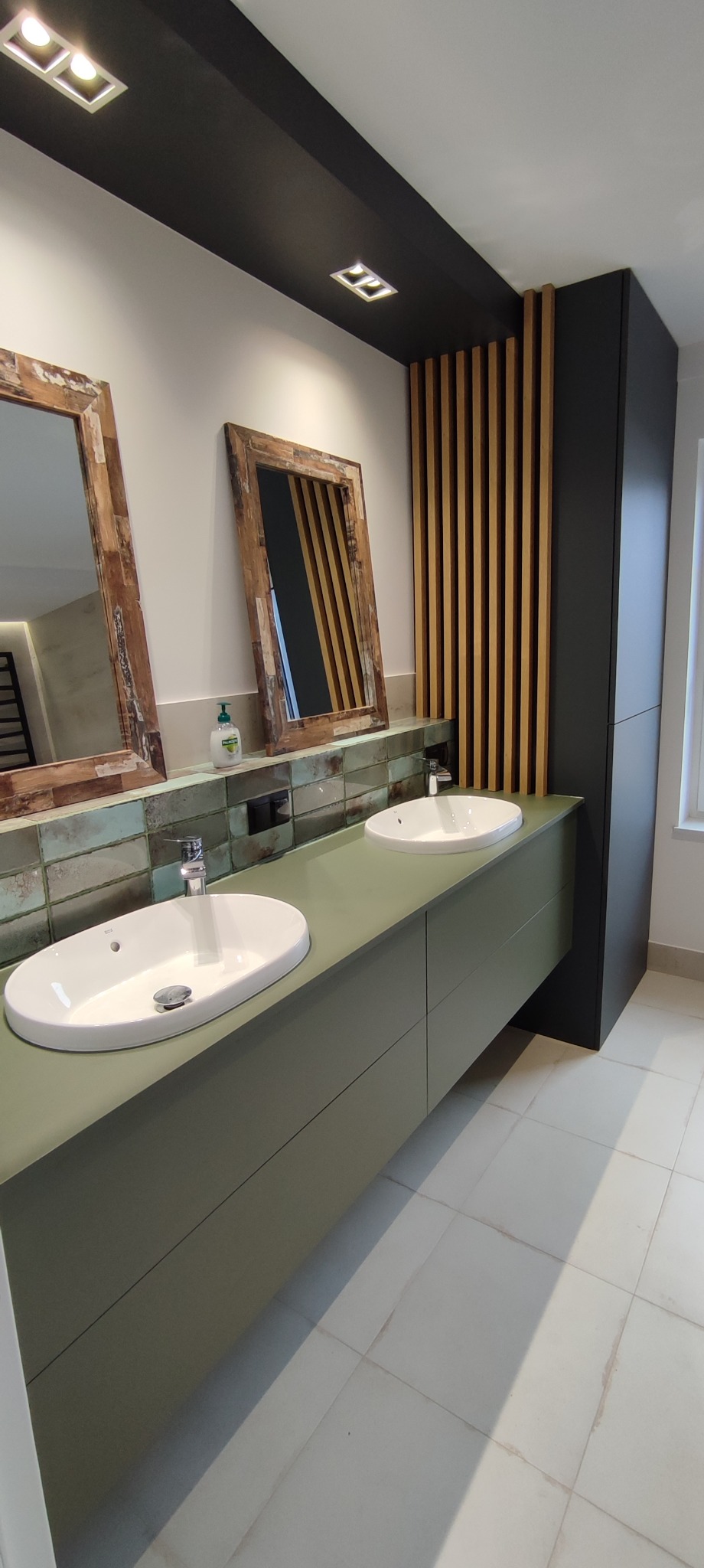 Nowoczesna łazienka z dwoma umywalkami nablatowymi, szafką w odcieniach zieleni, lustrami w drewnianych ramach i dekoracyjnymi drewnianymi lamelami obok czarnej szafki