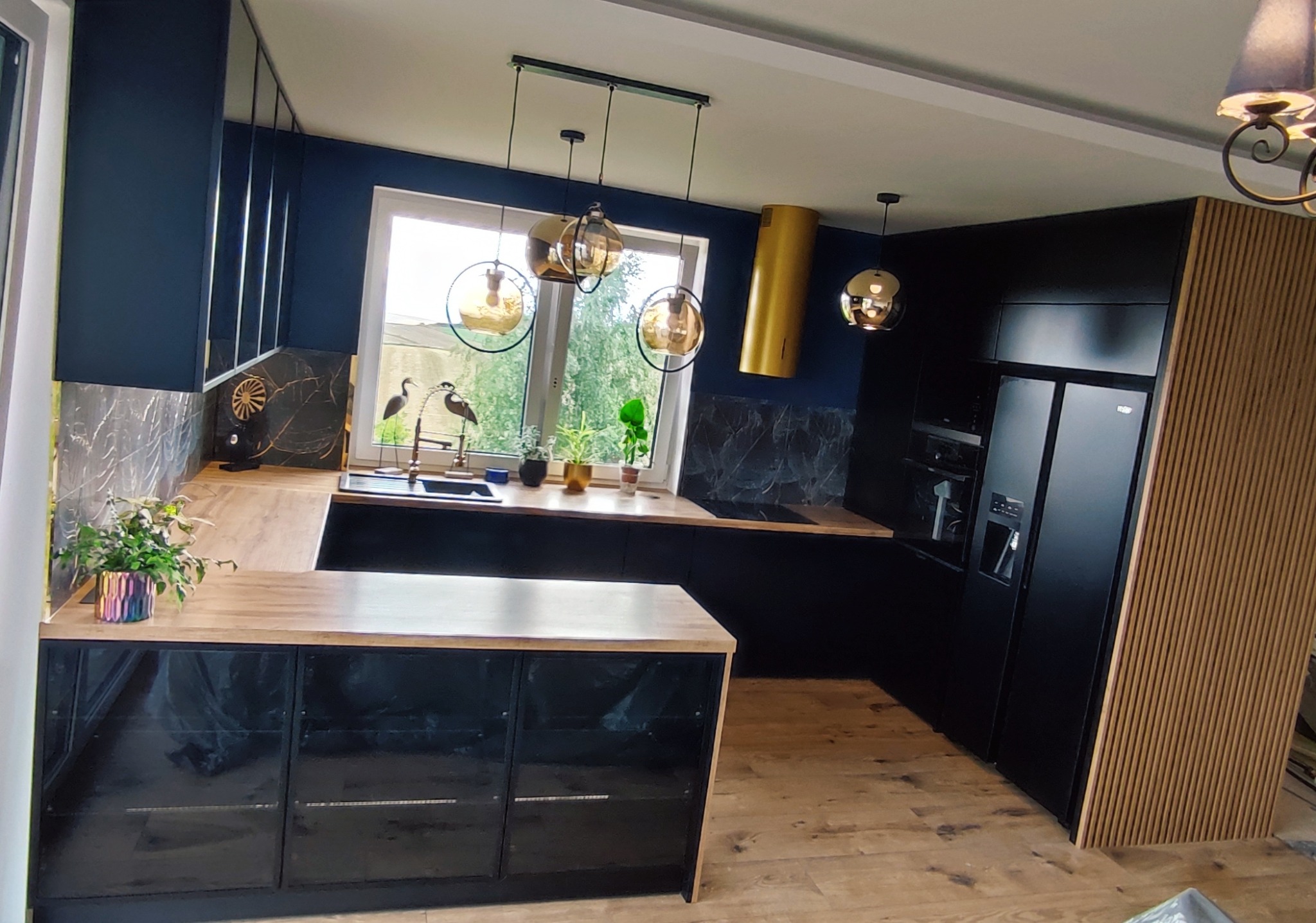 Nowoczesna kuchnia w kolorze granatowym z drewnianymi akcentami, wyspą z czarnymi frontami i blatem w kolorze jasnego drewna, okrągłymi lampami wiszącymi i złotym okapem.