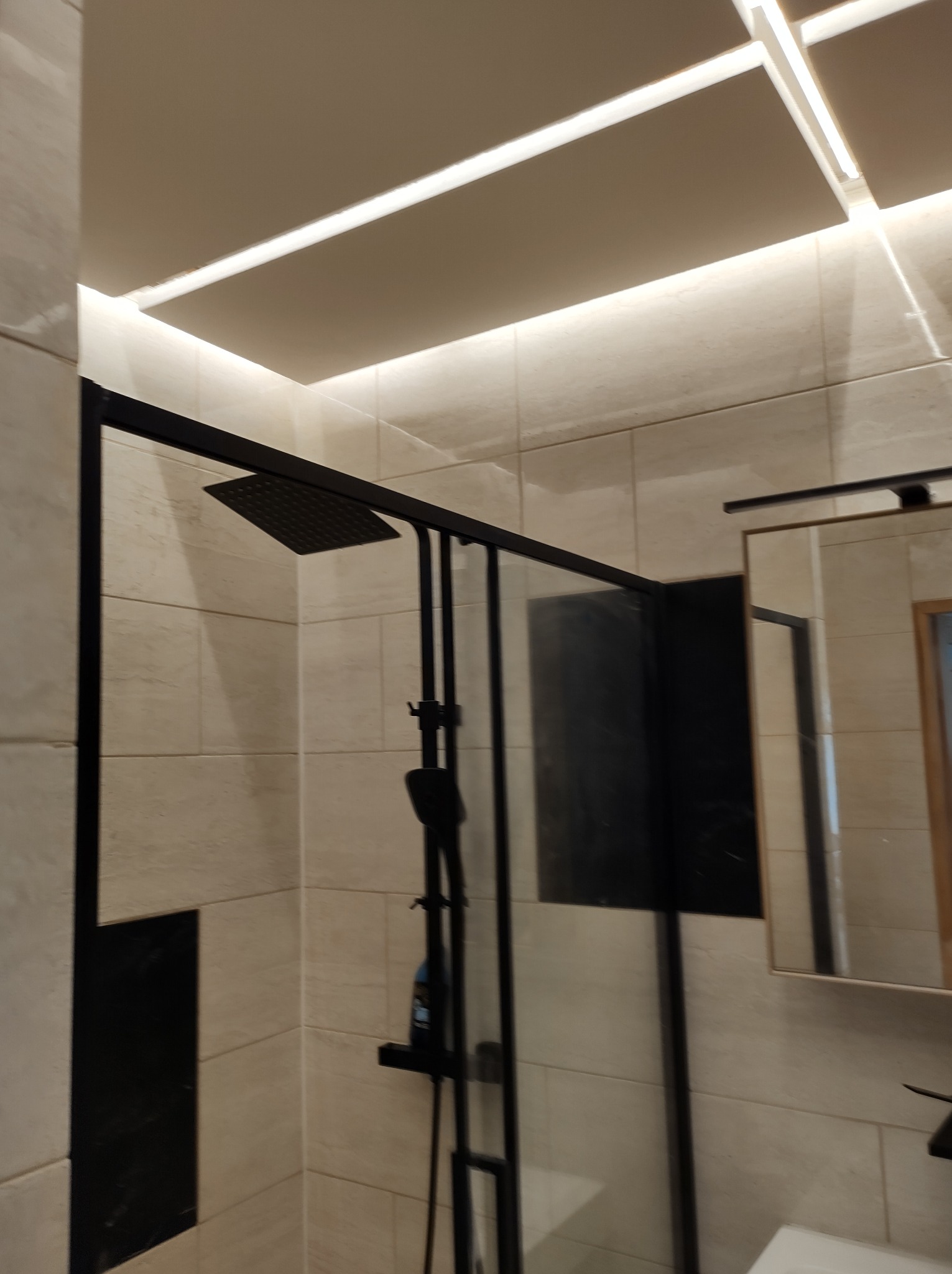 Nowoczesna kabina prysznicowa z czarnymi ramami i deszczownicą, jasne płytki ceramiczne na ścianach, oświetlenie LED w suficie podwieszanym.