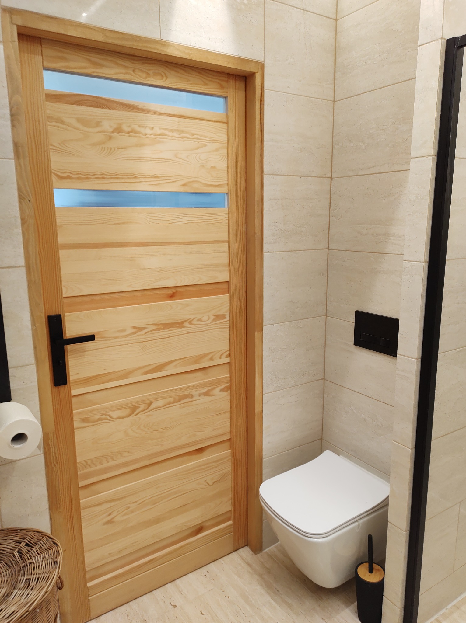 Wykończona łazienka z drewnianymi drzwiami z przeszkleniami, beżowe płytki na ścianach i podłodze, wisząca toaleta i czarne akcenty.