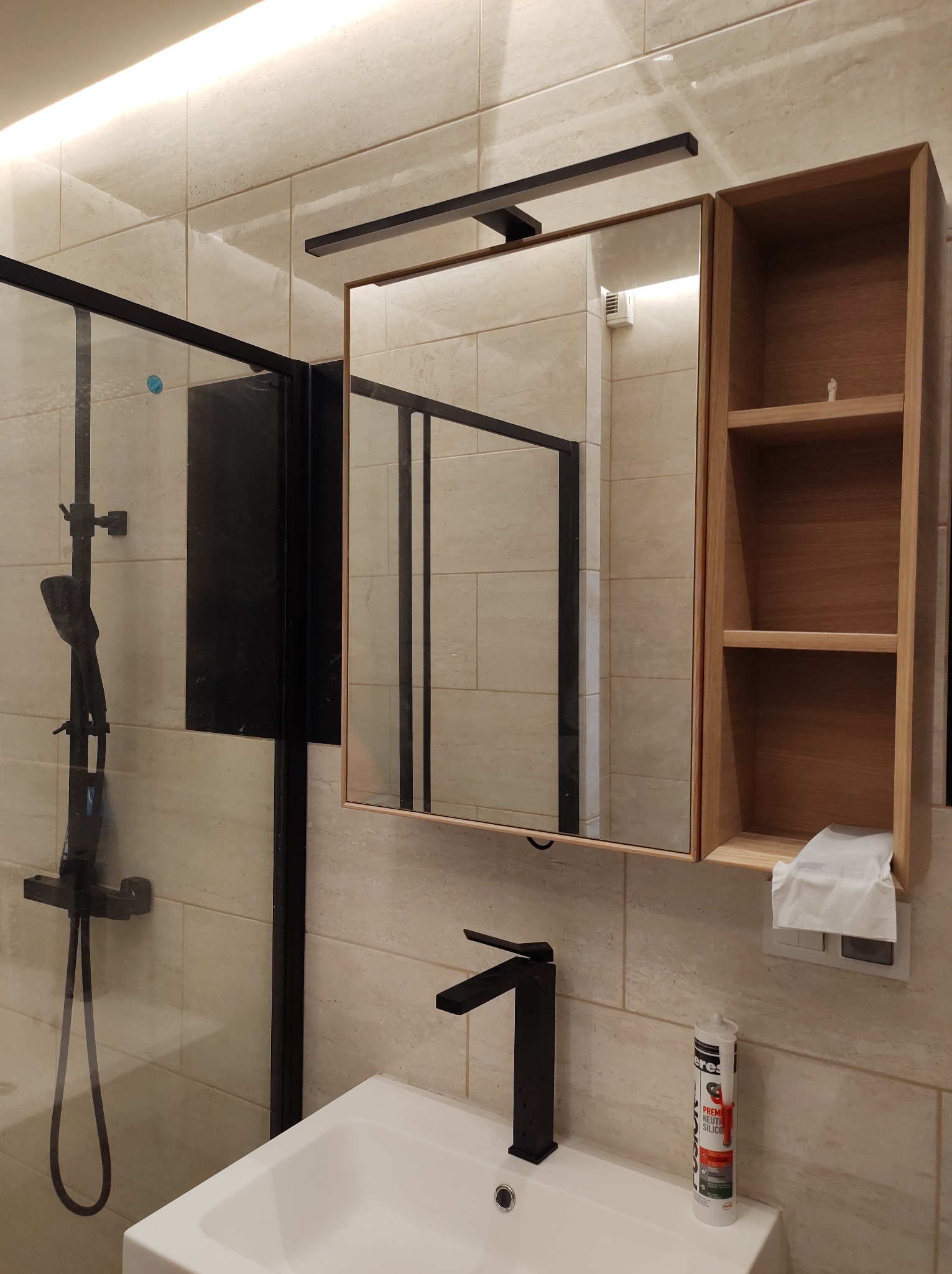 Nowoczesna łazienka z czarną armaturą: kabina prysznicowa typu walk-in, umywalka z baterią, lustro z oświetleniem i drewnianą szafką, kremowe płytki na ścianach, widoczna tuba silikonu na umywalce.