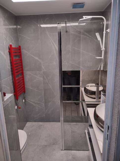 Nowoczesna łazienka z szarymi płytkami imitującymi marmur, kabiną prysznicową typu walk-in z chromowanymi profilami, czerwonym grzejnikiem drabinkowym i podwieszaną umywalką.