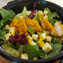 Sałatka z panierowanym kurczakiem, serem w kostkach i kukurydzą, podana w czarnym, plastikowym pojemniku na wynos.