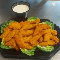 Ale dobre Karolina Krawczyńska - Porcja panierowanych filetów z kurczaka ułożonych na liściach sałaty, podana na czarnym, plastikowym talerzu obok pojemnika z sosem, na metalowym blacie.