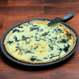 Quiche ze szpinakiem w okrągłej formie na drewnianym stole, z łopatką do ciasta, widok z góry.