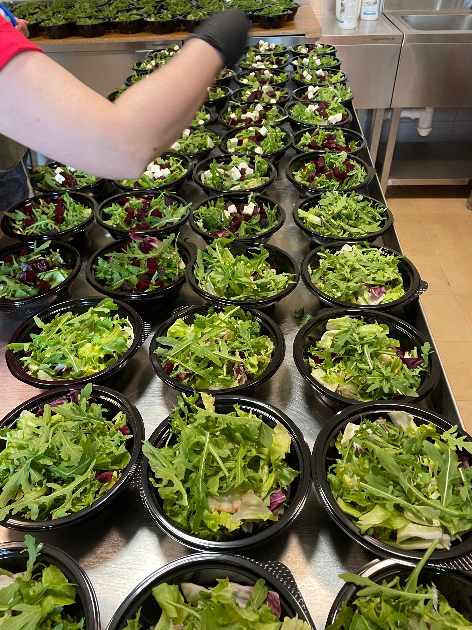 Przygotowywanie porcji sałatek z burakami i serem feta w czarnych miseczkach na metalowym stole, widoczna ręka w czarnej rękawiczce.