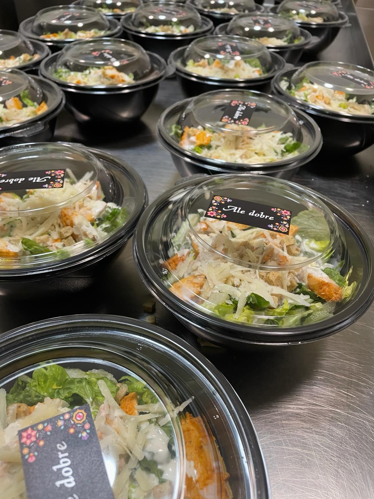Porcje sałatki Cezar w czarnych miseczkach z przezroczystymi pokrywkami, każda z naklejką 'Ale dobre' z kwiatowym motywem, ułożone rzędami na metalowym blacie.