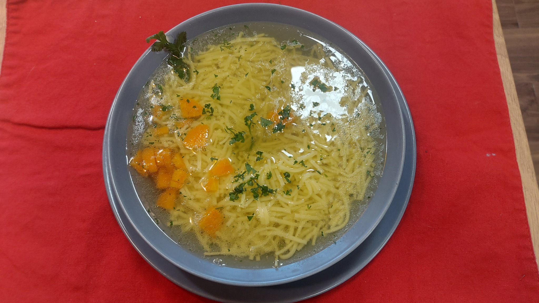 Talerz rosołu z makaronem i marchewką, udekorowany zieloną pietruszką, podany na czerwonym obrusie.