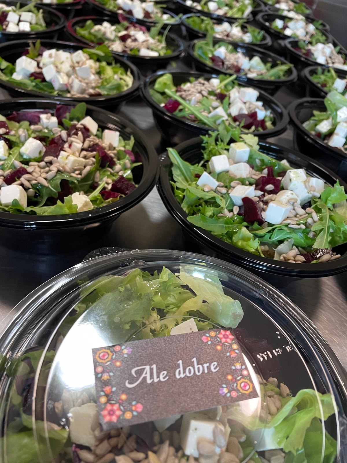 Rząd plastikowych misek z sałatką: zielenina, buraki, ser feta i nasiona słonecznika. Na jednym z pojemników naklejka z napisem 'Ale dobre' ozdobiona kwiatami.