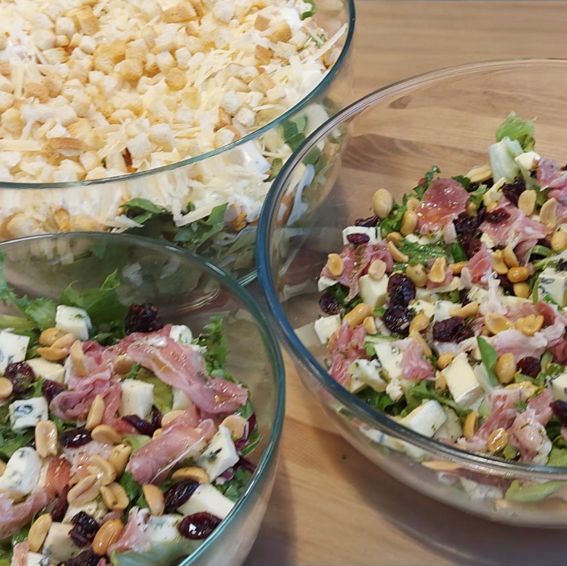 Trzy szklane miski z sałatkami: jedna z grzankami i serem, dwie z sałatą, serem pleśniowym, szynką parmeńską, orzechami i żurawiną, na drewnianym stole.