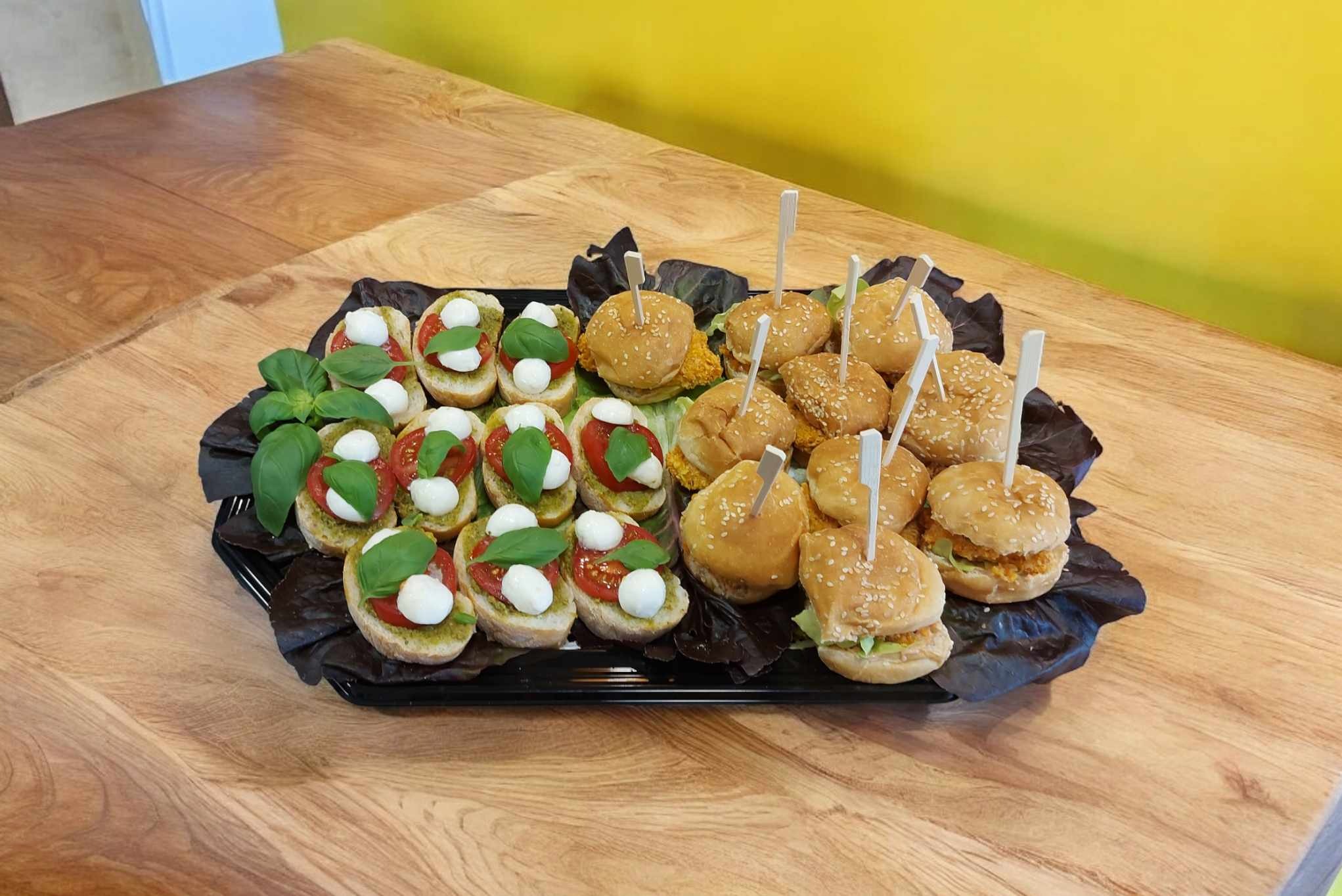 Elegancko podany catering: mini burgery z wykałaczkami i bruschetta caprese z bazylią na czarnym półmisku, na tle drewnianego stołu i żółtej ściany.