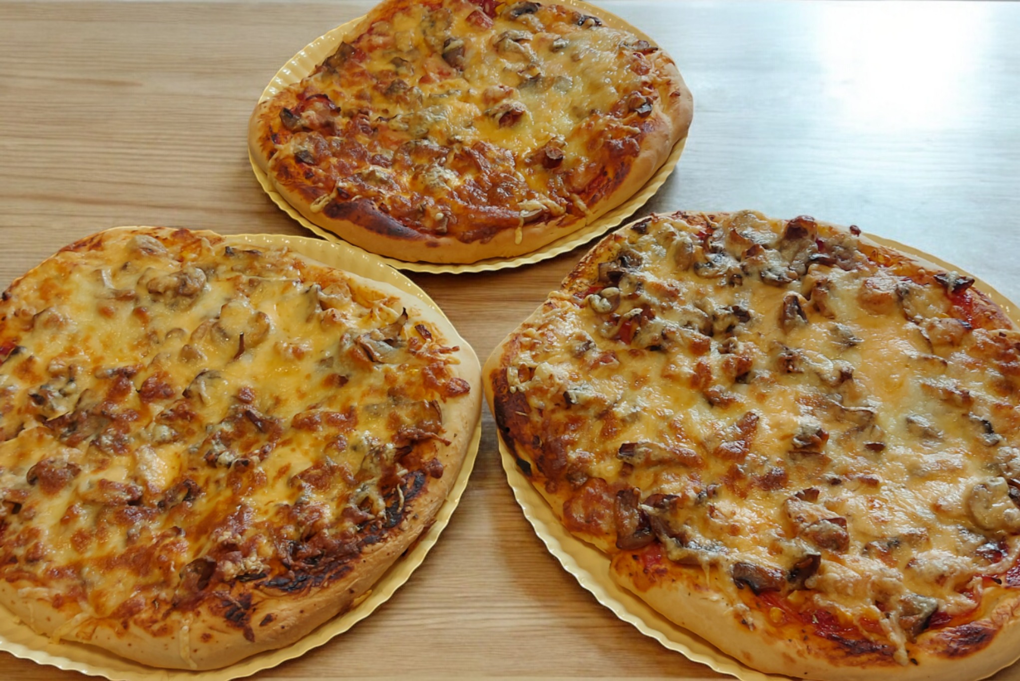 Trzy okrągłe pizze z pieczarkami i serem na tekturowych tackach, ułożone na jasnym drewnianym blacie.